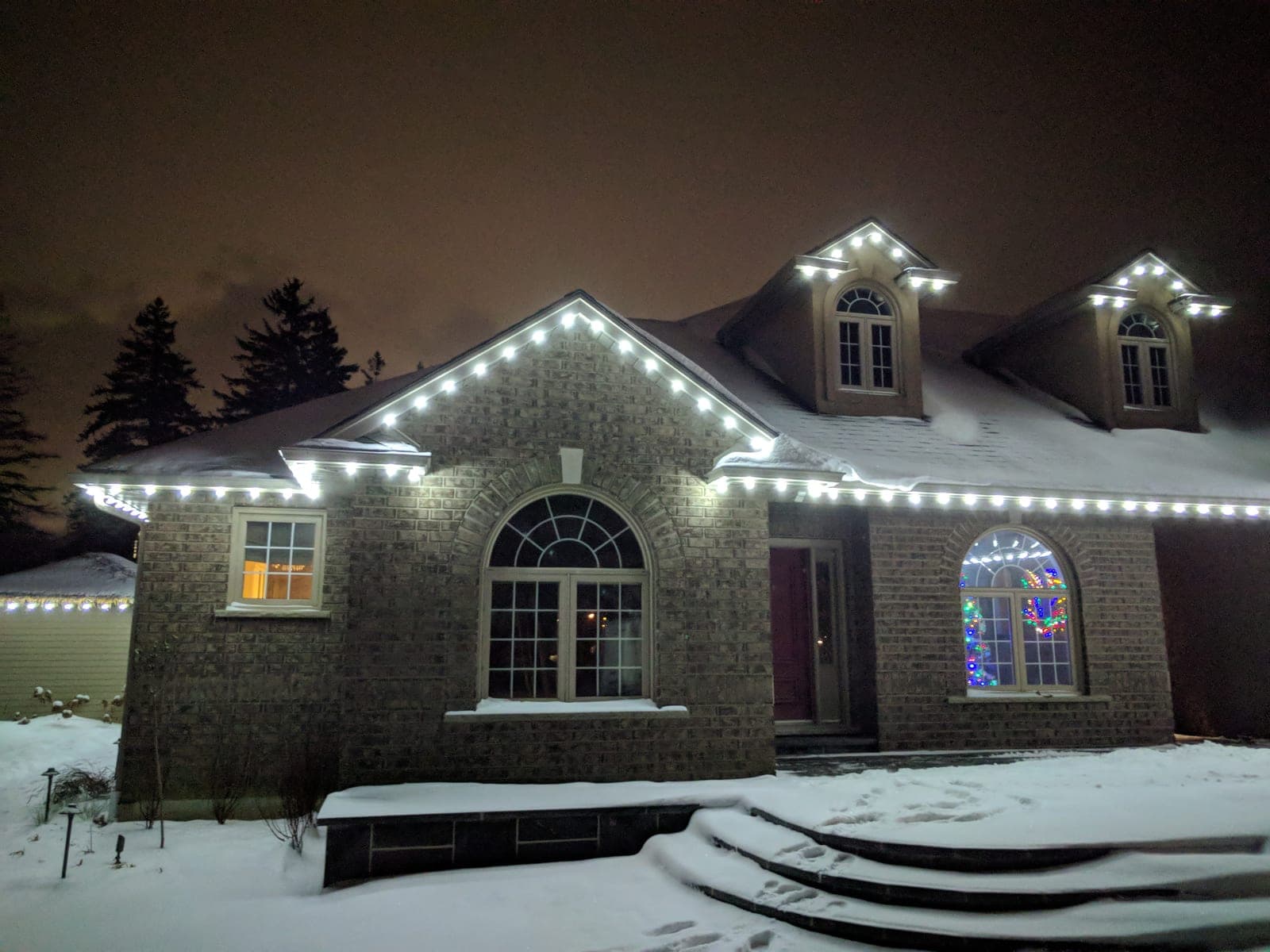 Front of a brick home lined with white roofline Christmas lights on a snowy winter night