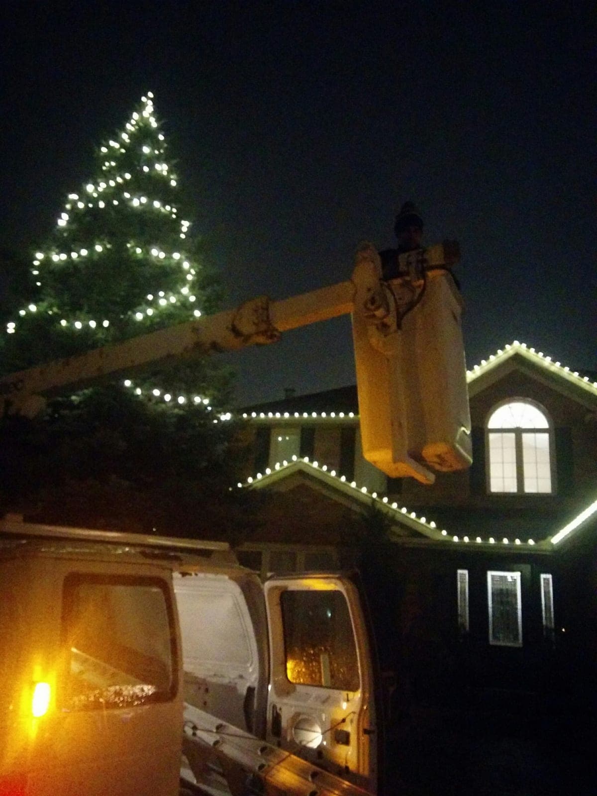 Bucket truck raising an installer to hang warm white Christmas lights along a home's roofline at night.