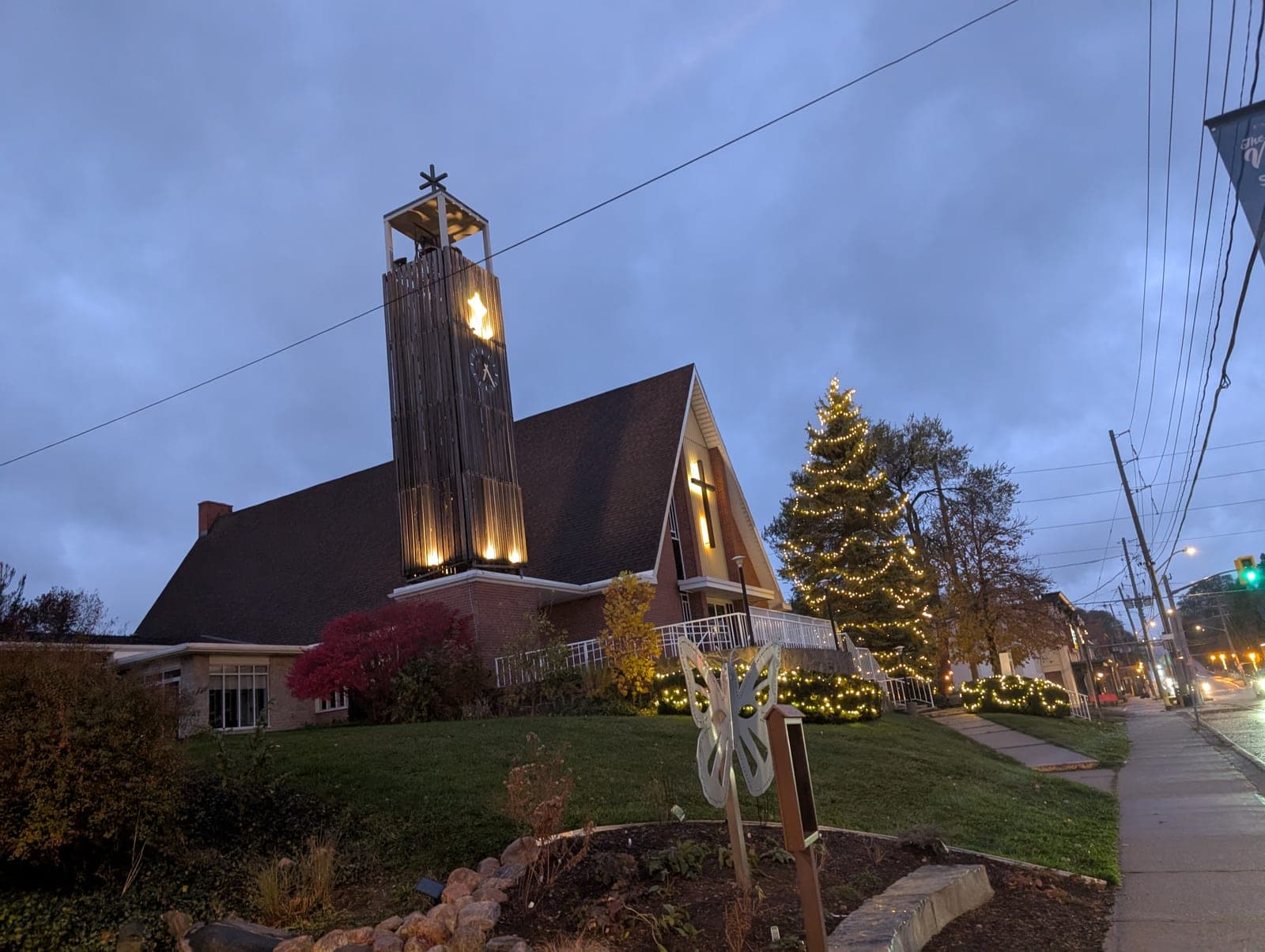 Church with an illuminated evergreen Christmas tree and light-wrapped shrubs along the sidewalk at dusk