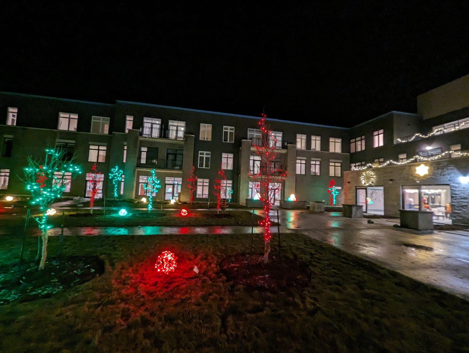 Nighttime holiday light display across a commercial property courtyard with colourful tree wraps and roofline lights.