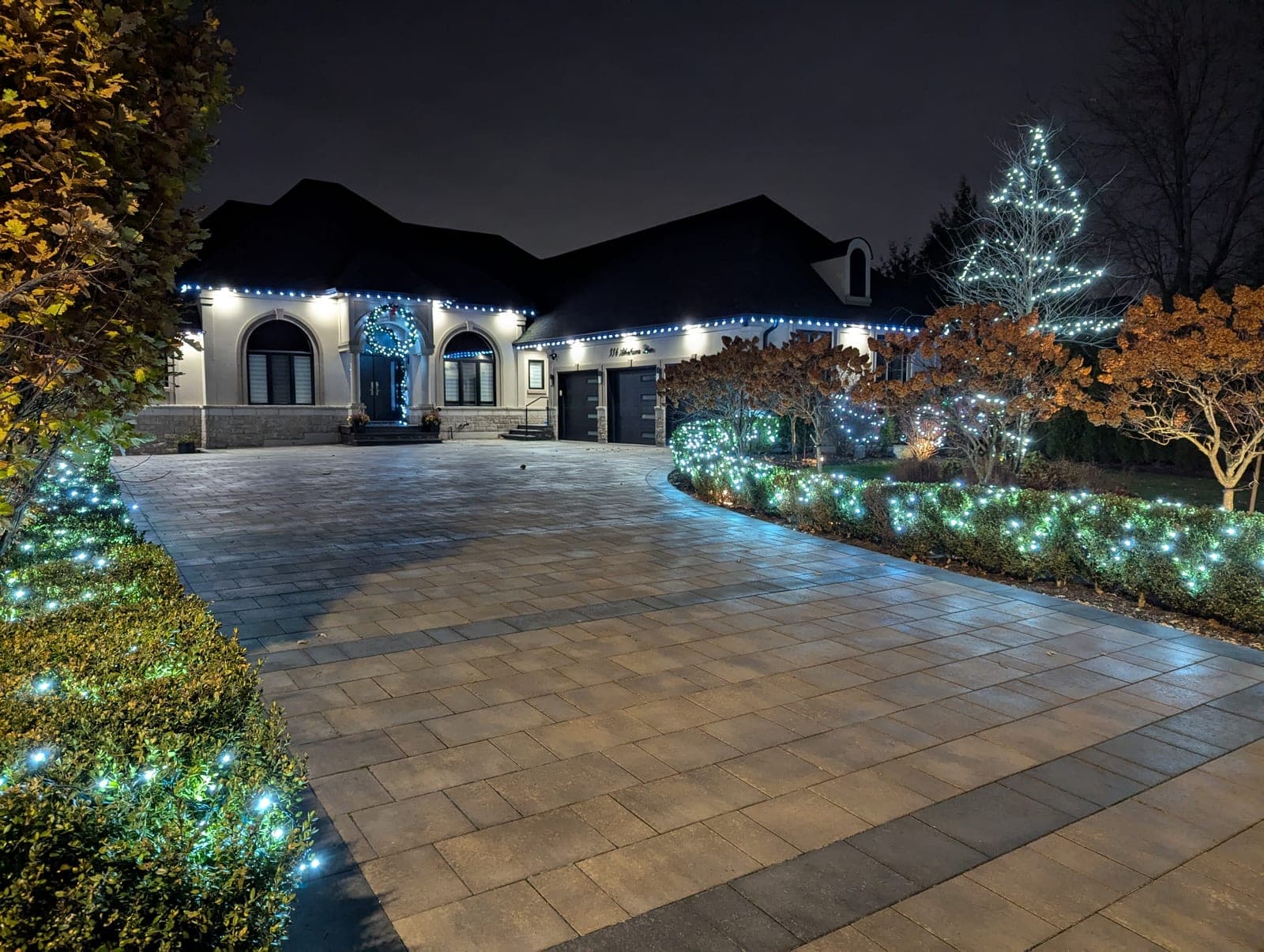 Estate home outlined in white Christmas lights with a light-wrapped tree and glowing shrub beds