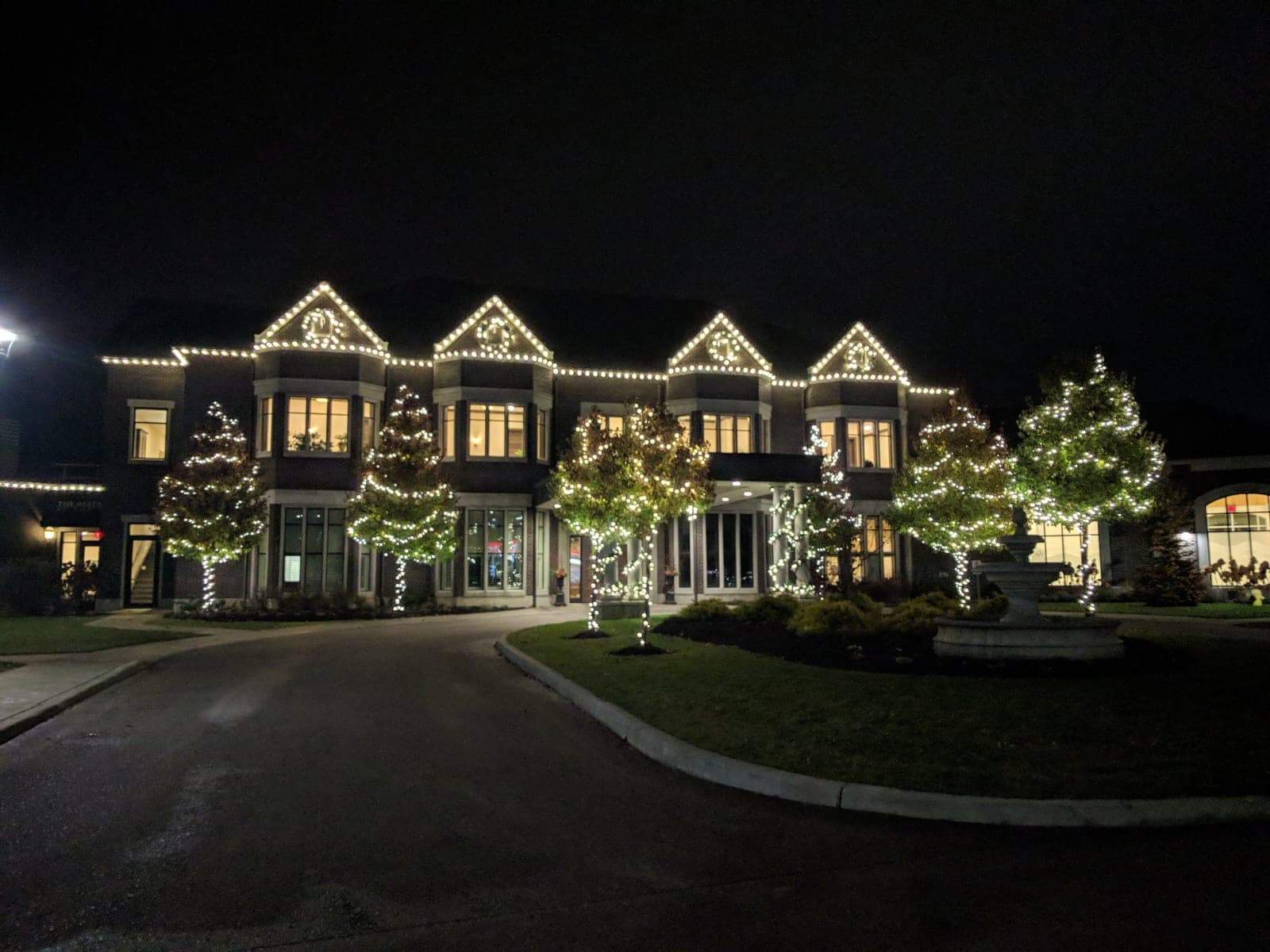 Grand estate venue lit with warm-white roofline lights and Christmas-wrapped trees at night.