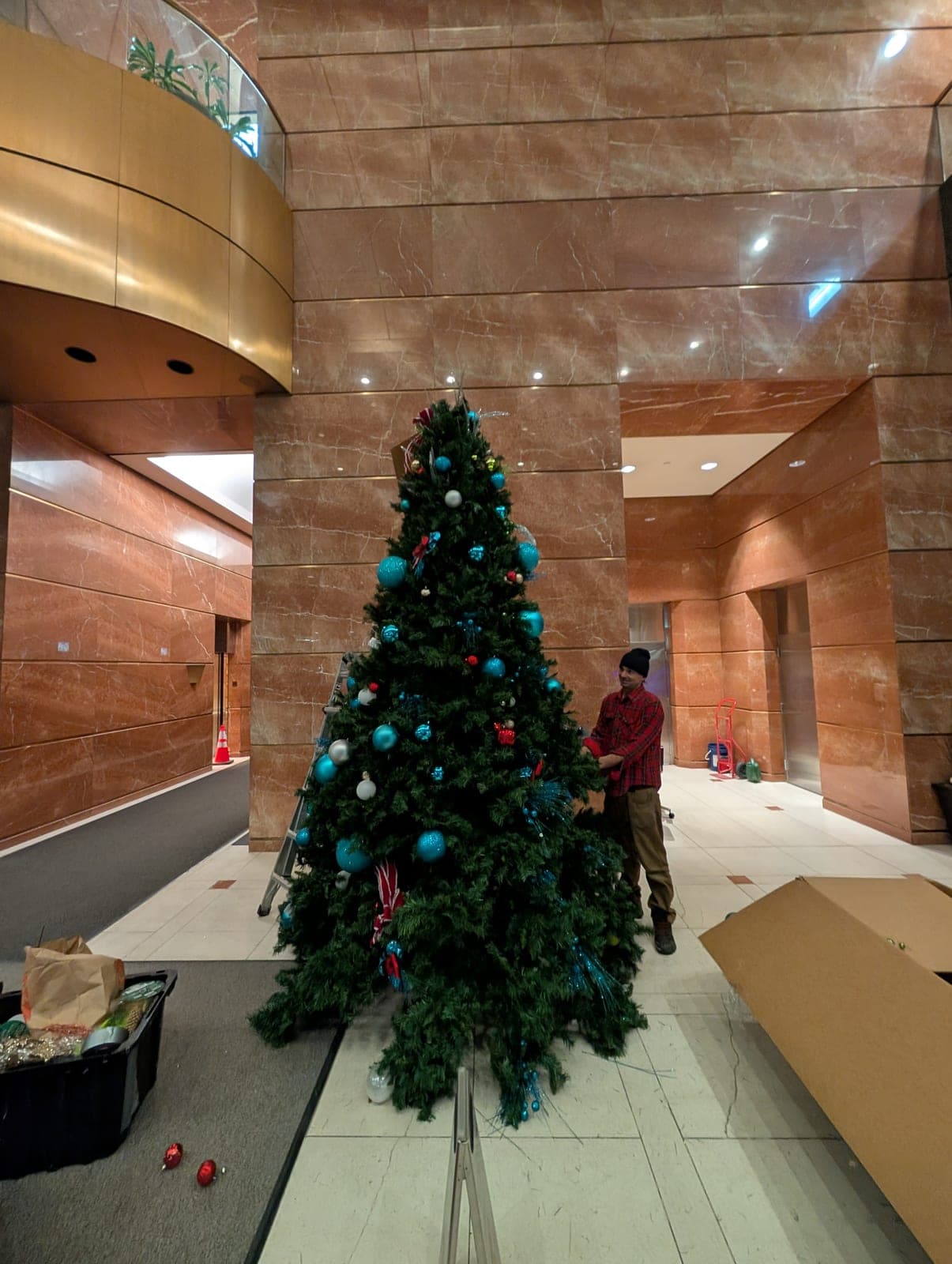 Worker decorating a large ornamented Christmas tree in a marble office tower atrium.