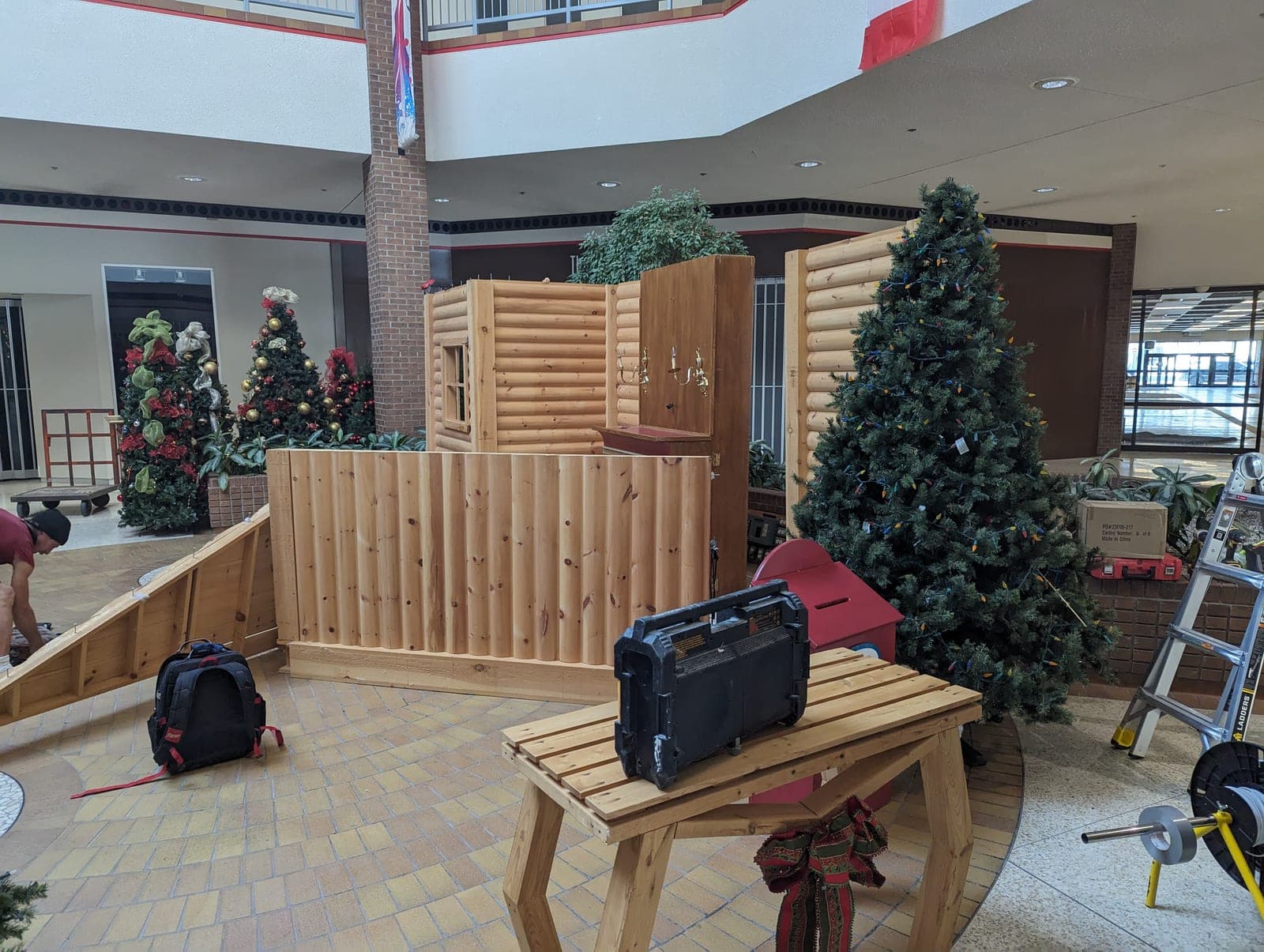 Installer setting up a log-cabin Santa Christmas village display in a shopping mall atrium with tools and ladder.