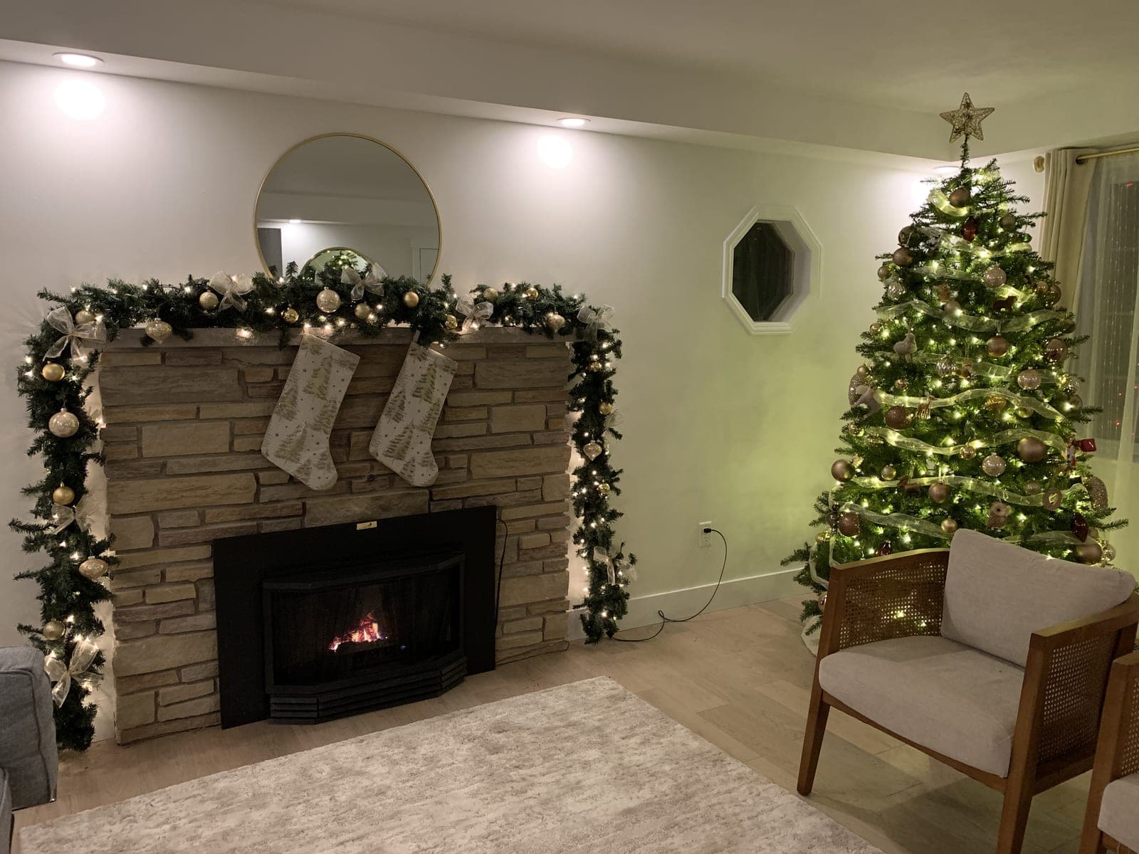 Living room with a lit Christmas tree beside a stone fireplace draped in warm-white garland lights and stockings