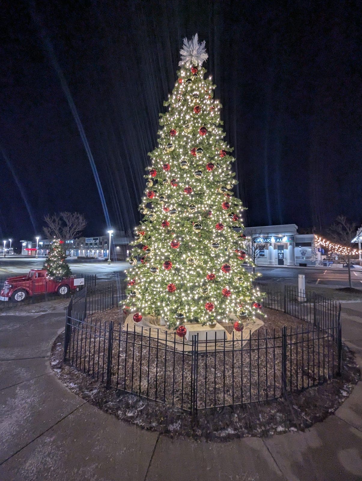 Two lit commercial Christmas trees and a vintage truck in a shopping plaza at night