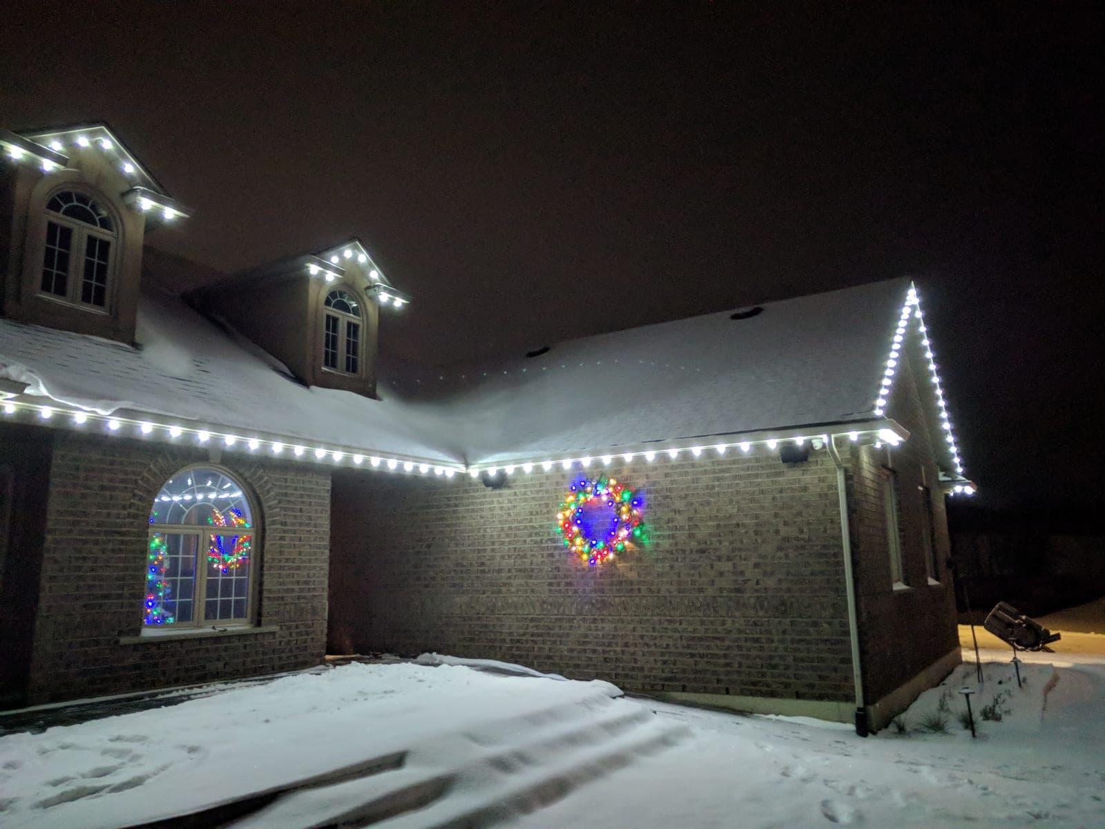 Brick home in fresh snow with white roofline lights and a multicolour lit wreath on the wall