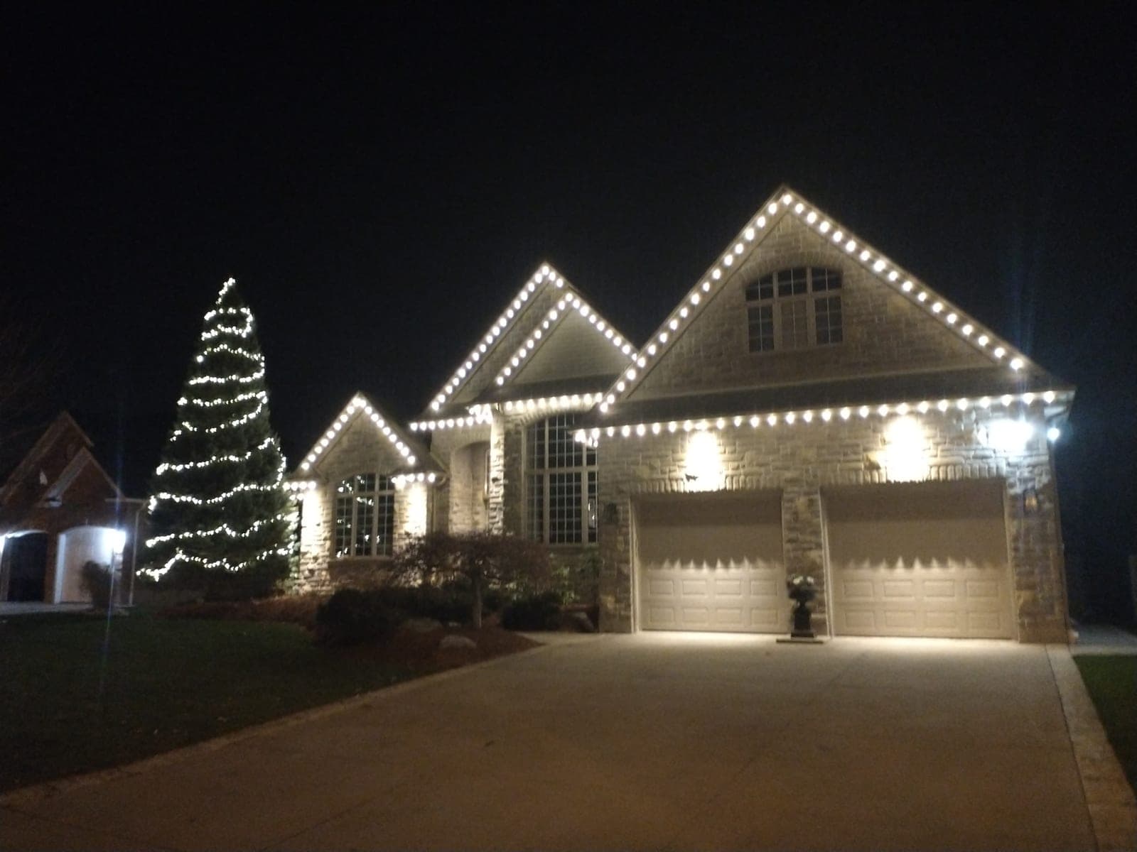 Two-storey stone home with crisp warm-white roofline Christmas lights and a lit evergreen