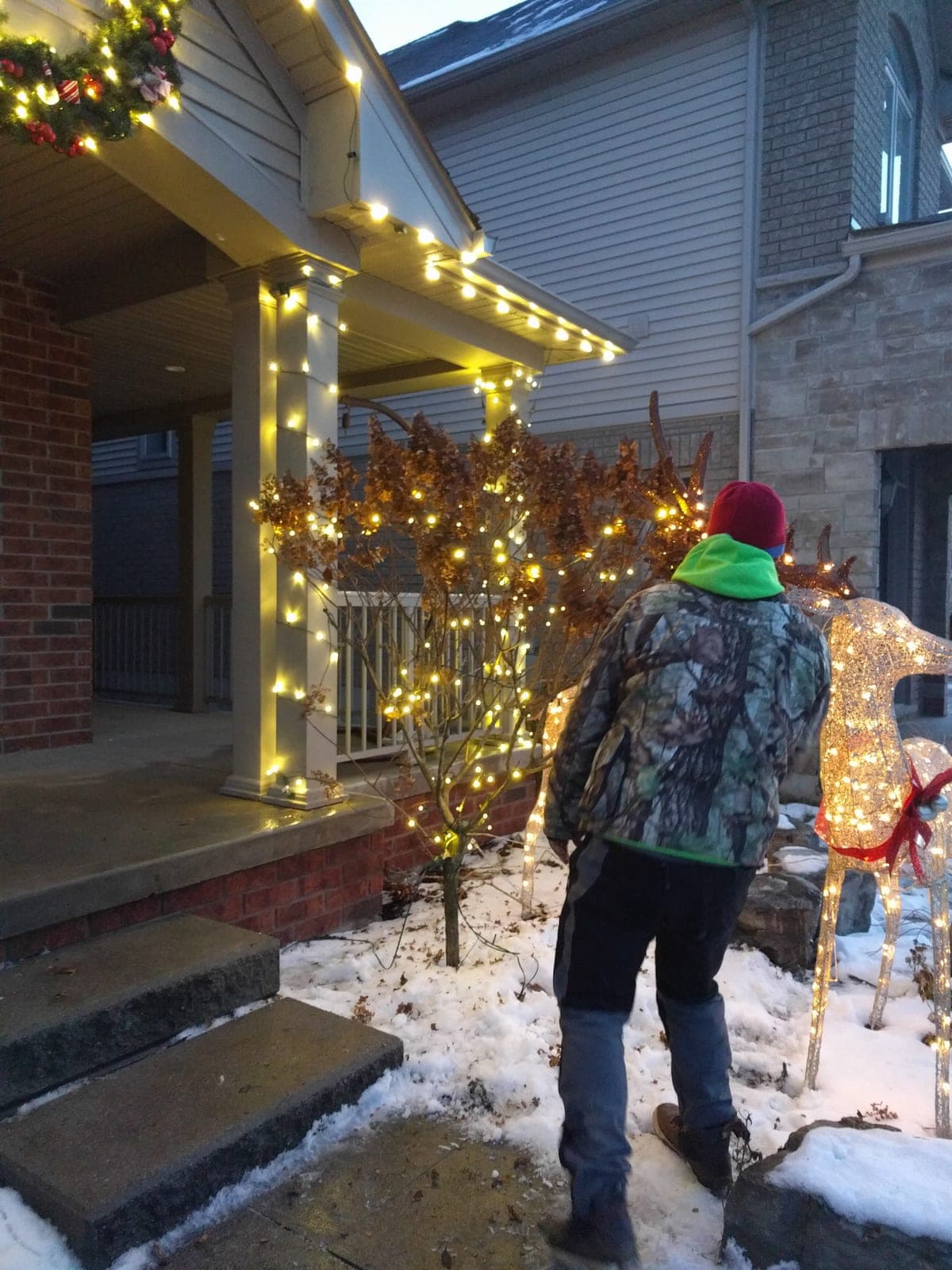 Decorated home entrance with warm white porch column lights, garland and a lit wire reindeer in the snow.