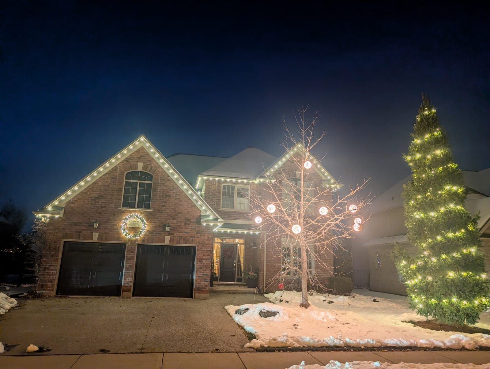 Two-storey brick home outlined in warm-white roofline Christmas lights with a lit evergreen tree