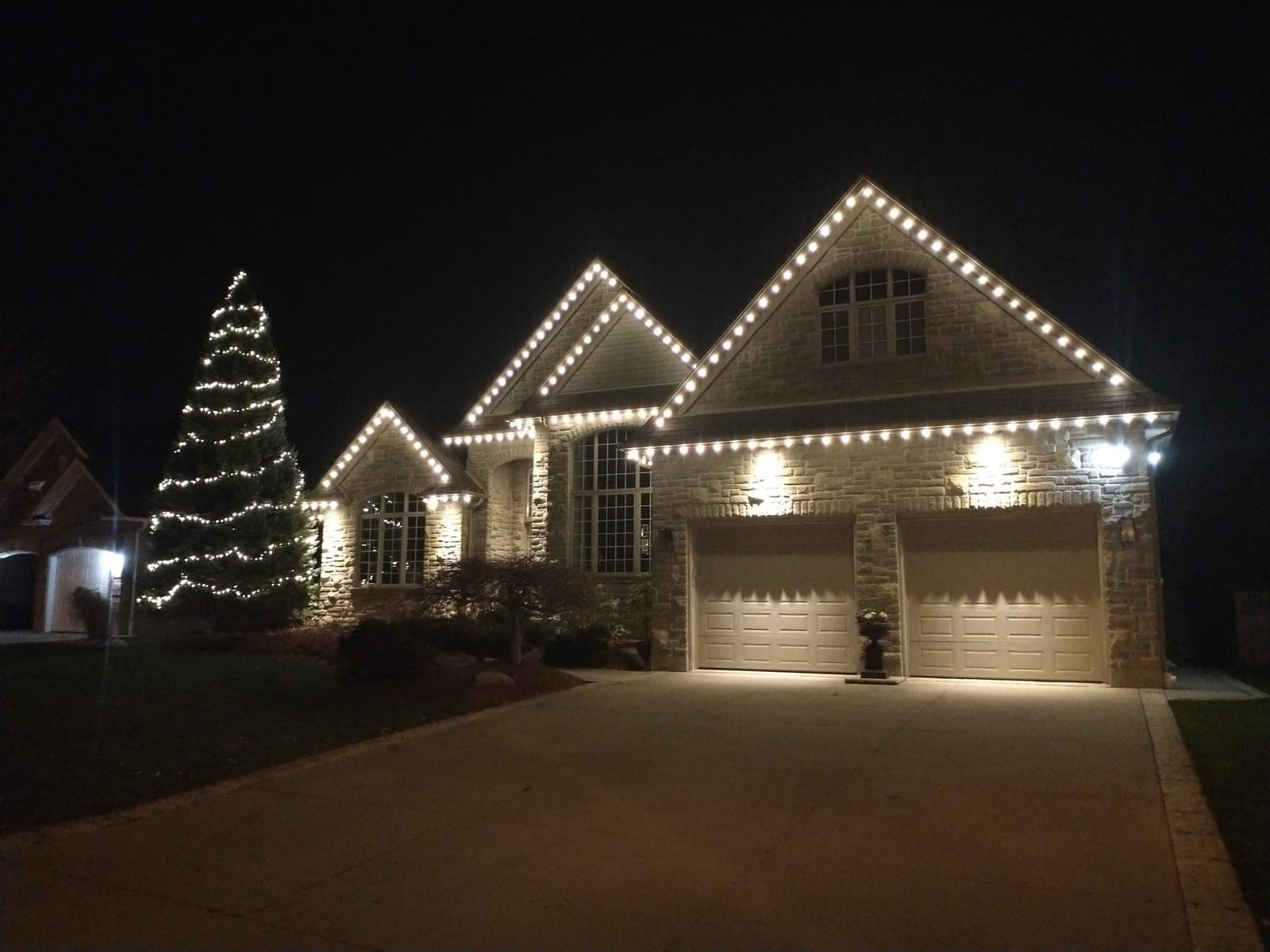 Stone home with warm-white roofline Christmas lights and a tall wrapped evergreen tree at night