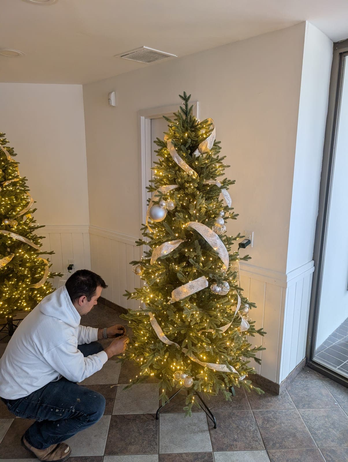 Installer kneeling to decorate a warm-white lit Christmas tree inside a commercial building.